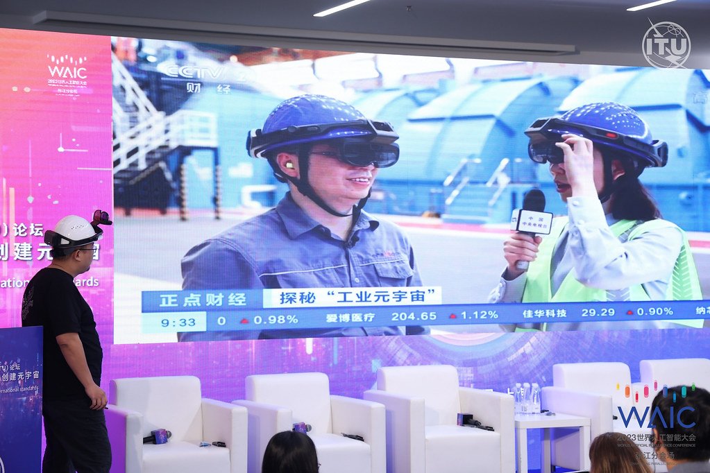 A man in a casual outfit observes a large display showing a woman in a high-visibility vest interviewing a man wearing a safety helmet, against the backdrop of an industrial setting. The screen is part of